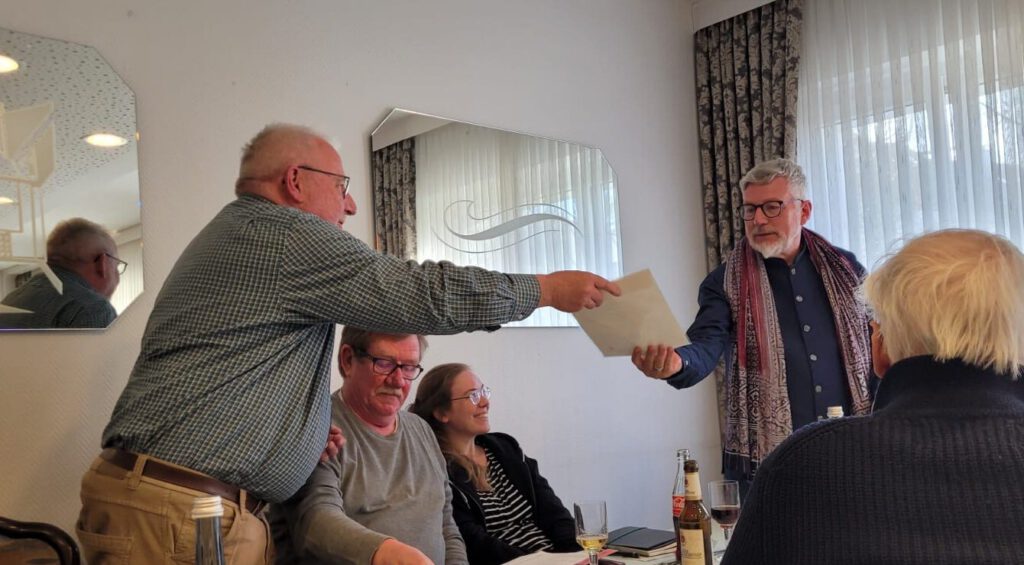 Foto der Jahreshauptversammlung 2025 der Kolpingsfamilie Velbert im Breuersaal: Überreichung der Urkunde zum Ehrenpräses. Auf dem Foto überreicht Michael Wolframm, Vorsitzender, die Urkunde an Peter Jansen. Beide halten ein Ende der Urkunde in der Hand. Zwischen den beiden sitzen zwei Vorstandsmitglieder. Aus Platzgründen musste die Überreichung über den Tisch erfolgen.