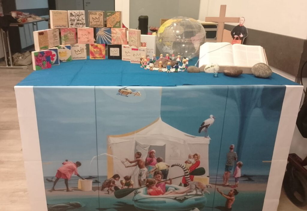 Das Foto ist im Rahmen des Bildungswochenendes der Kolpingsfamilie Velbert zum Thema Würde entstanden. Es zeigt den provisorischen Altar zum Wortgottesdienst. Auf dem Altar stehen - neben einer Bibel, einer Kleinen Figur von Adolph Kolping und dem Holzkreuz - Würdetafeln, die in der Nachmittagsaktion entstanden sind. Ebenfalls ist eine durchsichtige Weltkugel zu erkennen. Vor dem Tisch hängt das Misereor Hungertuch zur Fastenzeit 2025 /2026 „Liebe sei Tat“ von Konstanze Trommer.