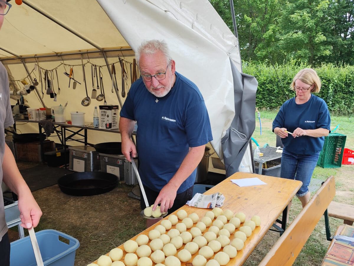 Zeltlager Kolping Velbert 2025 - Vor dem großen Küchenzelt steht eine Bierzeltgarnitur. Der Vorstand der Kolpingsfamilie Velbert bereitet mit dem Küchenteam das Essen vor. Auf dem Tisch liegen Knödel bereit.