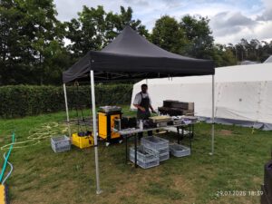 Zeltlager Kolping Velbert 2025 - Unter einem schwarzem Pavillion wird gekocht. Tim Vollmer bereitet Burger vor. Um ihn herum stehen diverse Kisten und Küchenutensilien. Er steht unter anderem neben einem Grill. Vor ihm auf dem Tisch stehen eine Friteuse und bereits ein bkech voller Pommes.