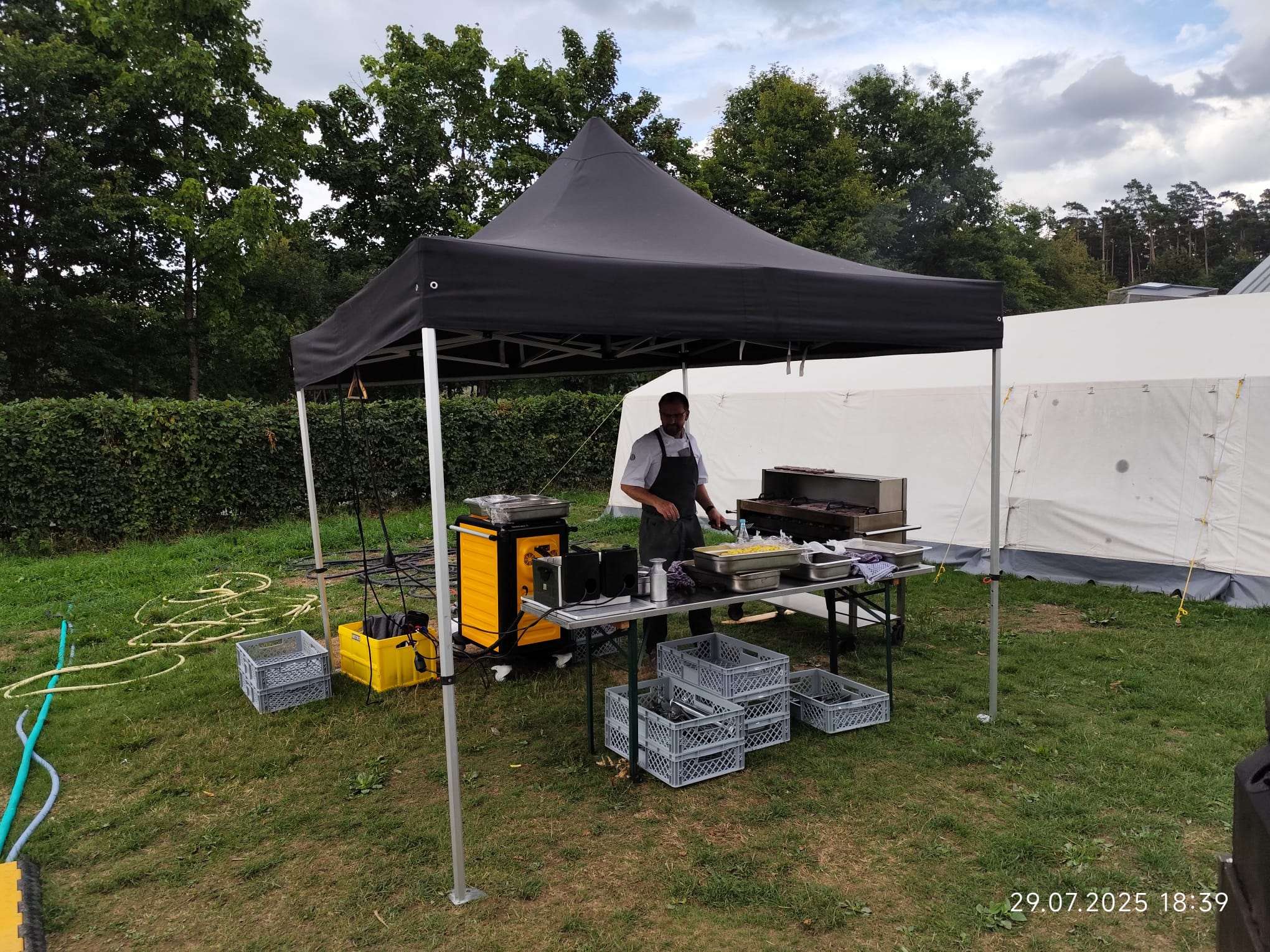 Zeltlager Kolping Velbert 2025 - Unter einem schwarzem Pavillion wird gekocht. Tim Vollmer bereitet Burger vor. Um ihn herum stehen diverse Kisten und Küchenutensilien. Er steht unter anderem neben einem Grill. Vor ihm auf dem Tisch stehen eine Friteuse und bereits ein bkech voller Pommes.