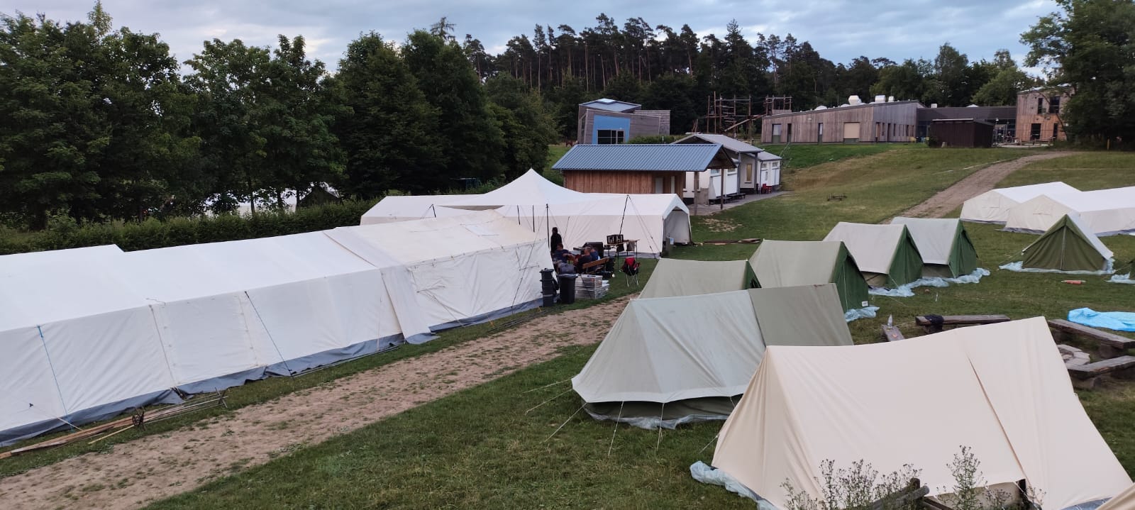 Zeltlager Kolping Velbert 2025 - Ein Foto vom Zeltlagerplatz. Mehrere weiße und kleinere grüne Zelte stehen auf einer Wiese, umgeben von Bäumen. Im Hinteren Bereich des Fotos ist das Haus des Zeltplatzes in Rüthen zu sehen sowie die Sanitärgebäude.