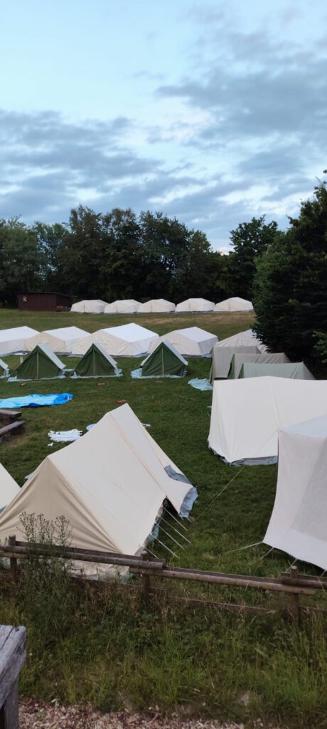 Zeltlager Kolping Velbert 2025 - Ein Foto vom Zeltlagerplatz. Mehrere weiße und kleinere grüne Zelte stehen auf einer Wiese, umgeben von Bäumen. Der Himmel ist an diesem Tag leicht bewölkt