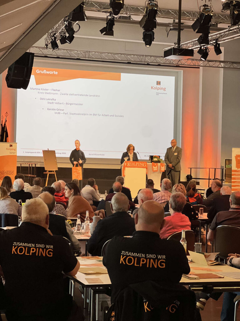 Foto Diözesanversammlung Diözesanverband Kolping Köln, im Hintergrund am Redepult Kerstin Griese, davor Teilnehmende an Tischen. Im Vordergrund zwei Herren der KF Velbert mit bestickten Shirts "Zusammen sind wir Kolping"