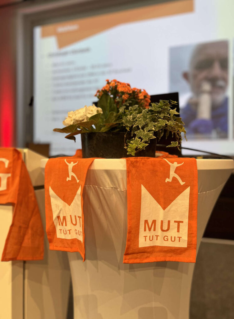Foto Diözesanversammlung Diözesanverband Kolping Köln, Impression Dekoration der Pulte auf der Bühne mit Blumen und Läufern in Orange "Mut tut Gut"