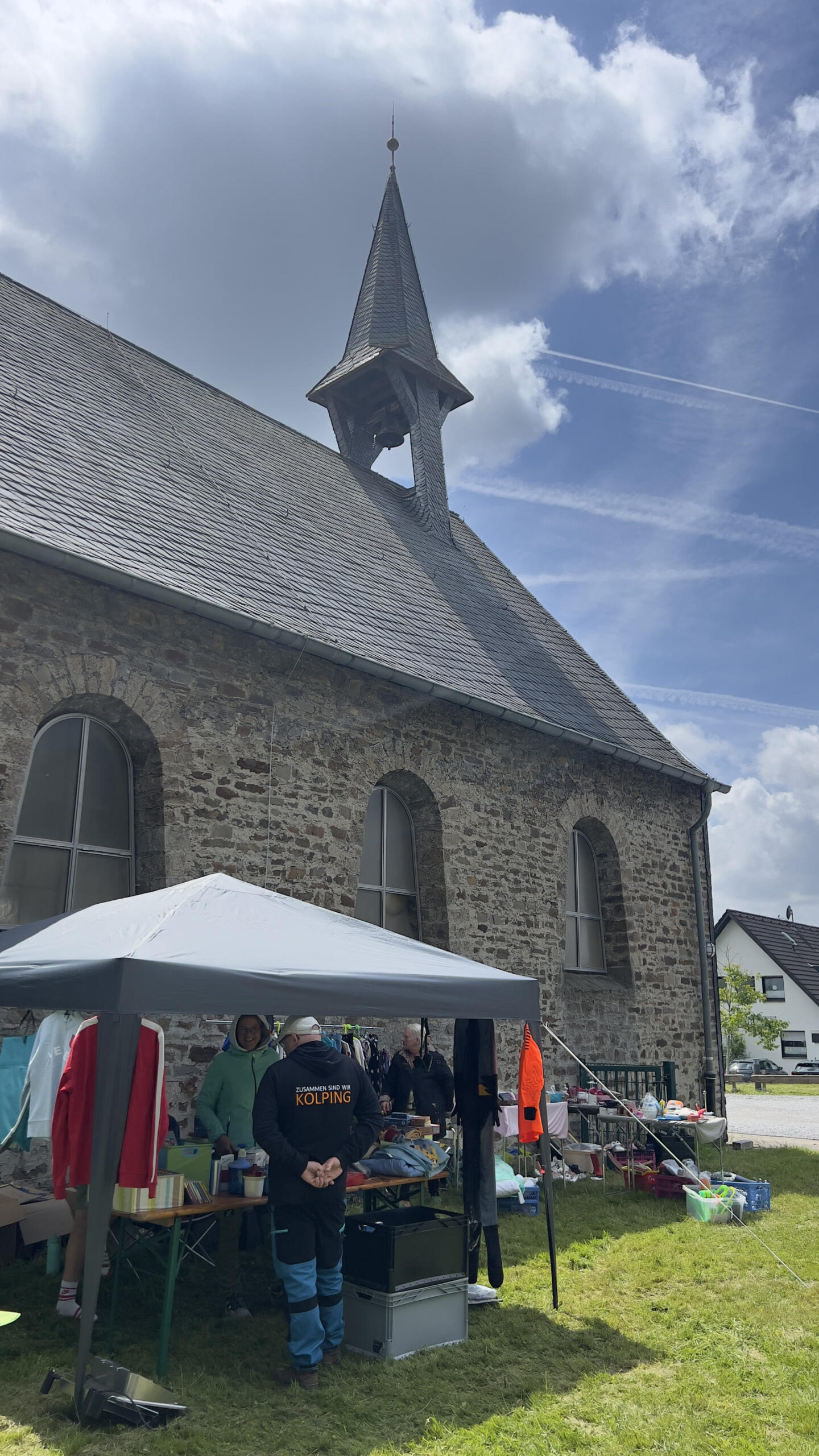 Foto vom Trödelmarkt an St. Nikolaus. Ein Stand mit Pavillion steht auf der Wiese direkt an der Außenwand der Kapelle. Die Kapelle ist aus Backstein und hat einen kleinen süßen Klockenturm.